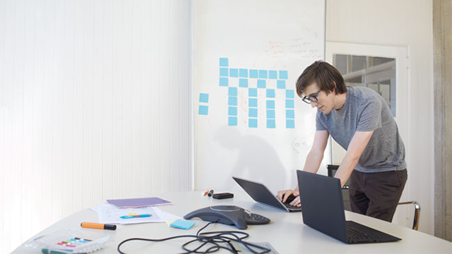 Person standing and leaning over a desk working on one laptop with another laptop beside it.